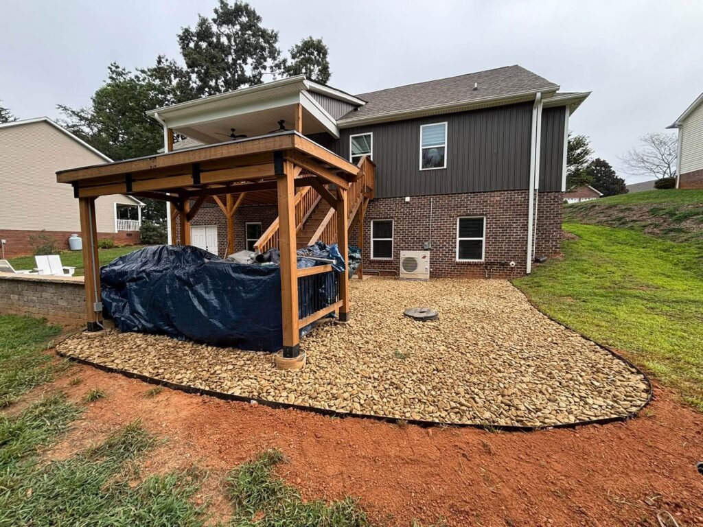 Backyard with a timber pavilion, tarp-covered furniture, and a curved river rock gravel bed edged with metal behind a brick