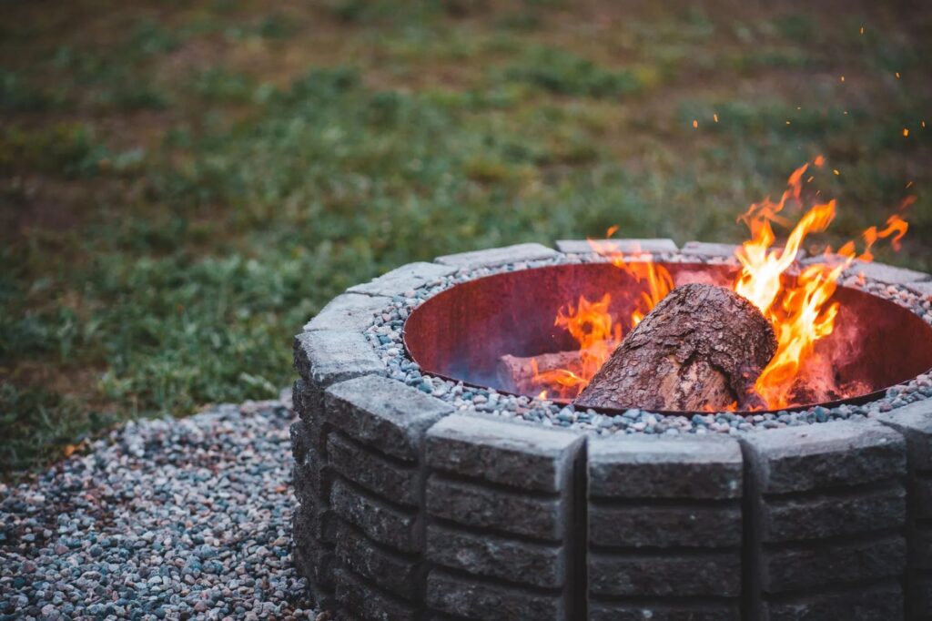 Close-up of a round concrete block fire pit with a rusted steel insert and burning logs, set on a pea gravel pad beside a