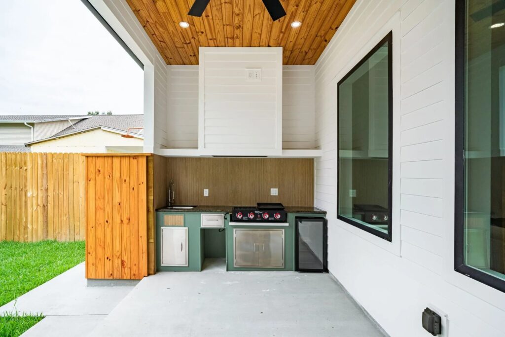 Covered patio outdoor kitchen with green cabinets, stainless doors, flat-top griddle, sink and beverage fridge under a wood