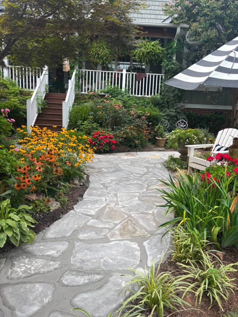 Irregular gray flagstone walkway winding between flower beds with coneflowers and roses toward a porch with white railings