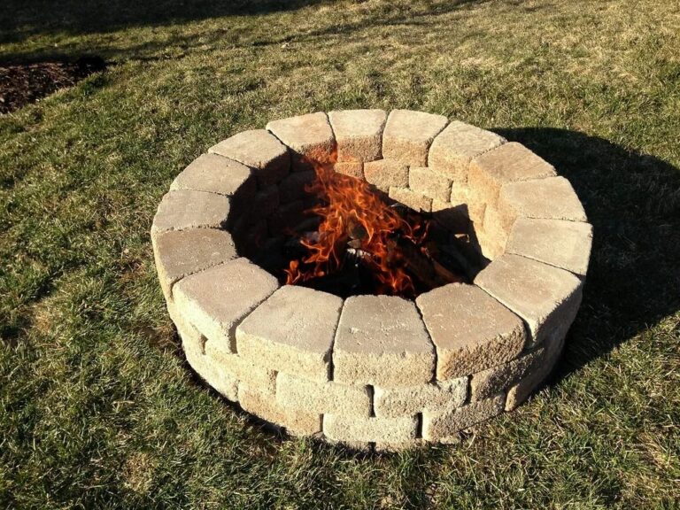 Round fire pit built from stacked tan concrete blocks with a wood fire burning inside, set on a grass lawn.