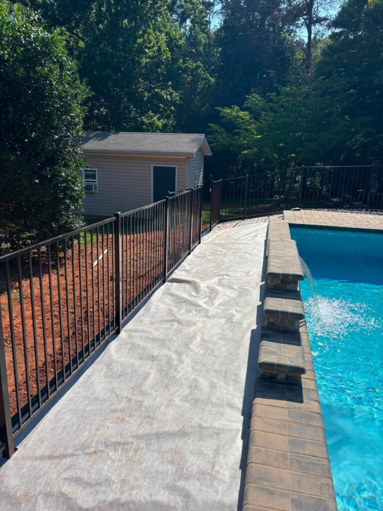 Landscape fabric laid along a pool deck beside a black aluminum fence, with a stone raised wall and waterfall spouts into the