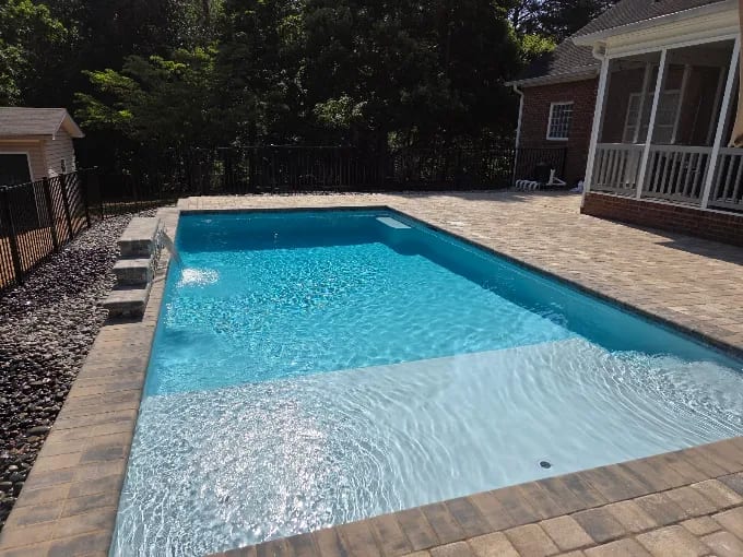 Rectangular in-ground pool with a shallow tanning ledge, tumbled paver deck, river rock border, and black aluminum fence