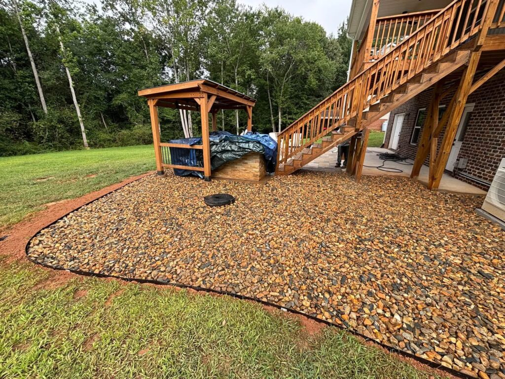 Curved river rock gravel bed with black edging beside a wooden deck staircase and pergola behind a brick house in Hickory, NC