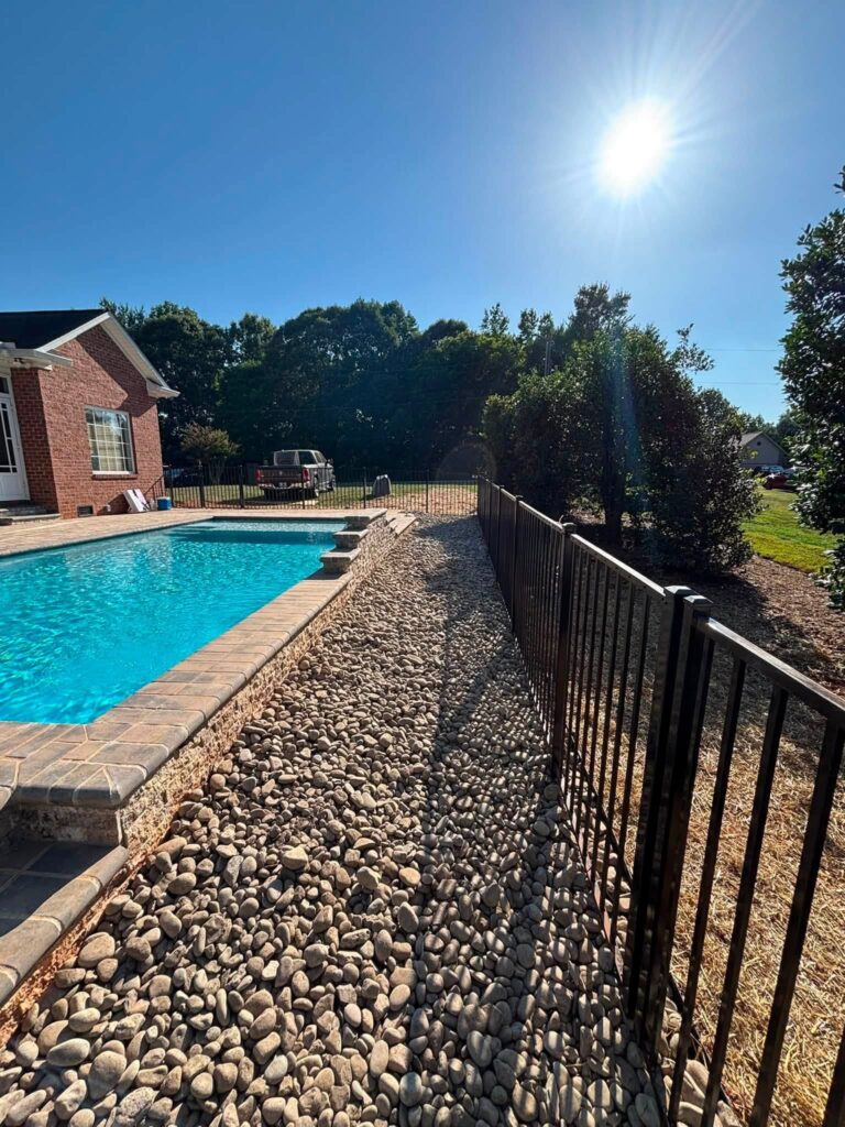 River rock drainage border between a paver pool deck and black aluminum fence beside a brick home on a sunny day.
