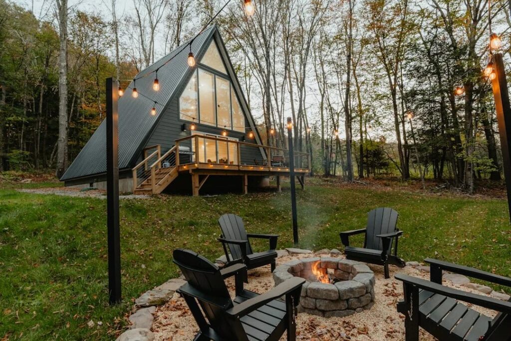 Round stacked stone fire pit with four black Adirondack chairs on gravel in front of a dark A-frame cabin with string lights