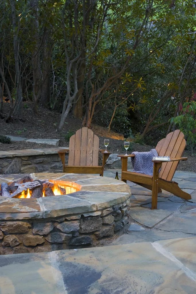 Round stacked stone fire pit with burning logs on a flagstone patio, two wooden Adirondack chairs and wine glasses behind it